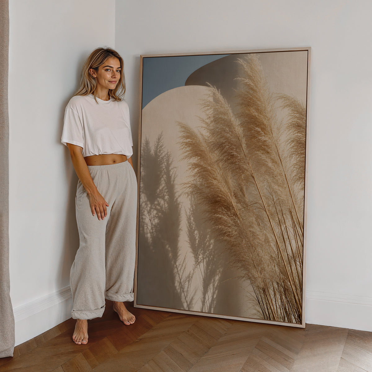 Motiv mit Pampasgräsern und ihren Schatten vor geschwungenen sandfarbenen Wänden unter blauem Himmel, als großes gerahmtes Wandbild neben einer Frau.