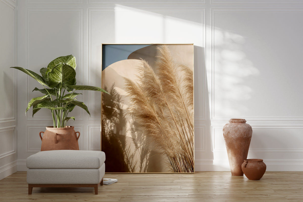 Motiv mit Pampasgräsern und ihren Schatten vor geschwungenen sandfarbenen Wänden unter blauem Himmel, als gerahmtes Poster neben Sessel und Zimmerpflanze.