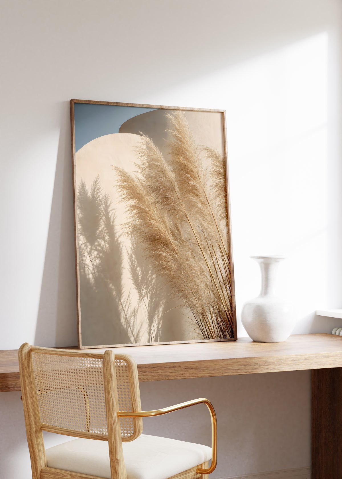 Gerahmtes Poster mit Pampasgräsern und ihren Schatten vor geschwungenen sandfarbenen Wänden unter blauem Himmel, auf einem Holztisch neben einer weißen Vase.