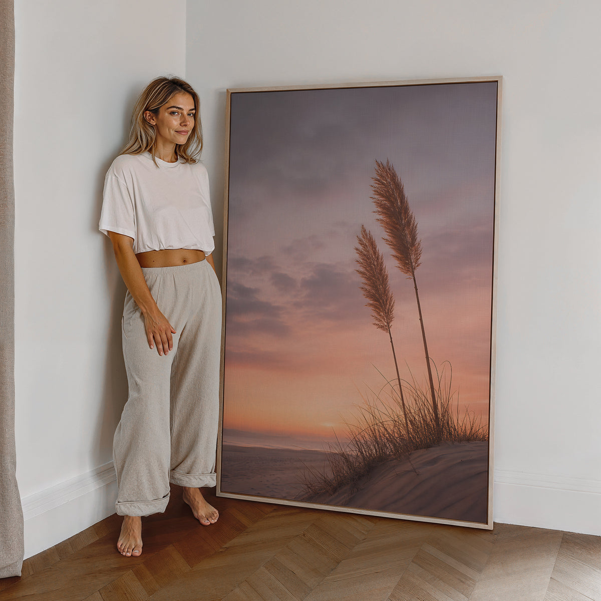 Motiv mit zwei Pampaswedeln auf einer Düne vor einem rosa-orangefarbenen Abendhimmel, als großes gerahmtes Wandbild neben einer Frau.