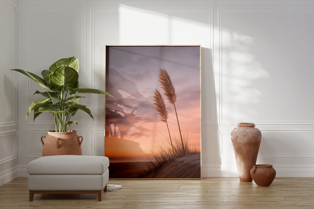 Motiv mit zwei Pampaswedeln auf einer Düne vor einem rosa-orangefarbenen Abendhimmel, als gerahmtes Poster neben Sessel und Zimmerpflanze.