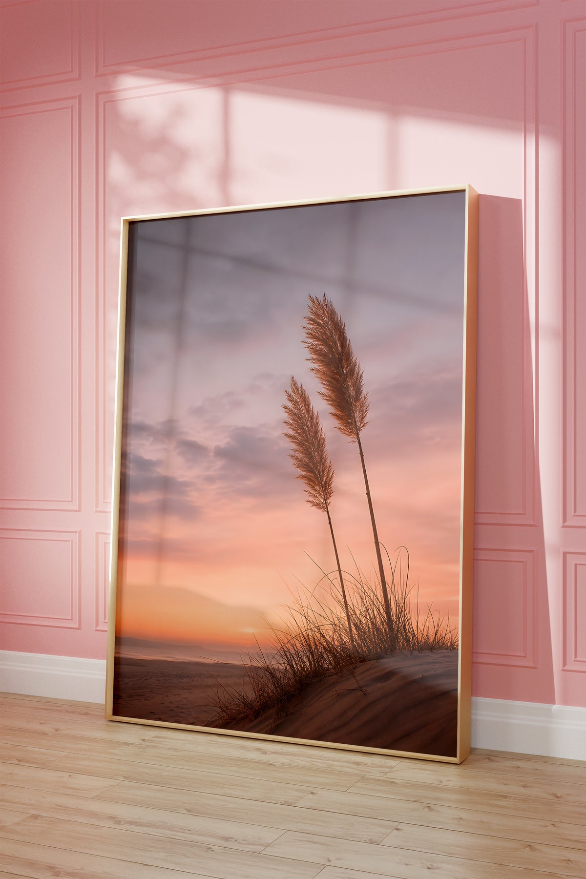 Motiv mit zwei Pampaswedeln auf einer Düne vor einem rosa-orangefarbenen Abendhimmel, als gerahmtes Poster vor einer rosafarbenen Wand.