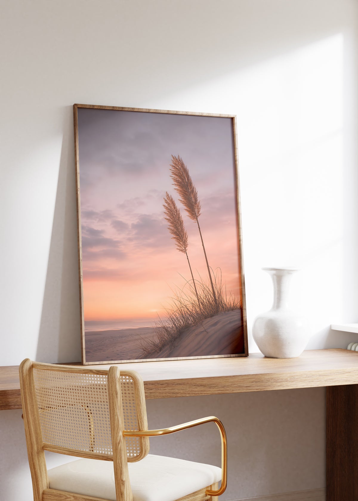 Gerahmtes Poster mit zwei Pampaswedeln auf einer Düne vor einem rosa-orangefarbenen Abendhimmel, auf einem Holztisch neben einer weißen Vase.
