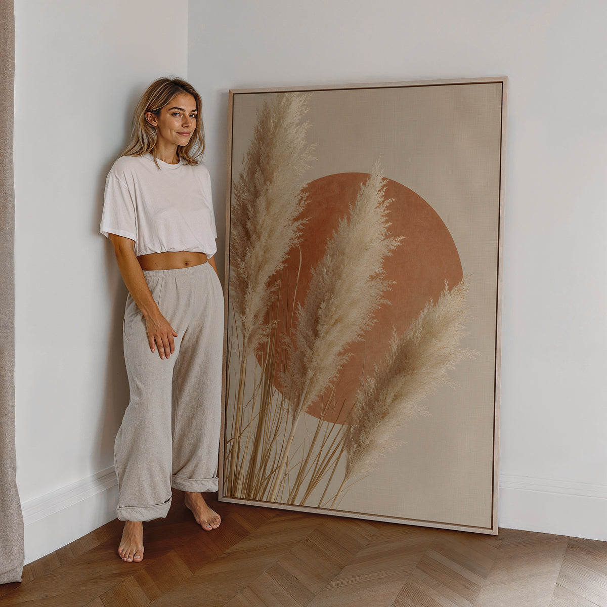 Motiv mit hellen Pampaswedeln vor einem großen terrakottafarbenen Kreis, als großes gerahmtes Wandbild neben einer Frau.