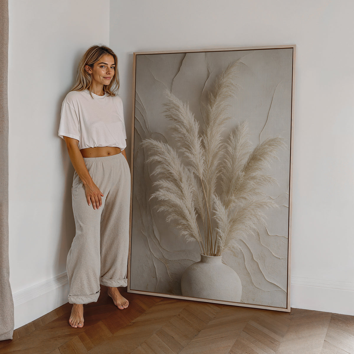 Motiv mit einem hellen Pampasbouquet in einer weißen Vase vor einer reliefartig strukturierten Wand, als großes gerahmtes Wandbild neben einer Frau.