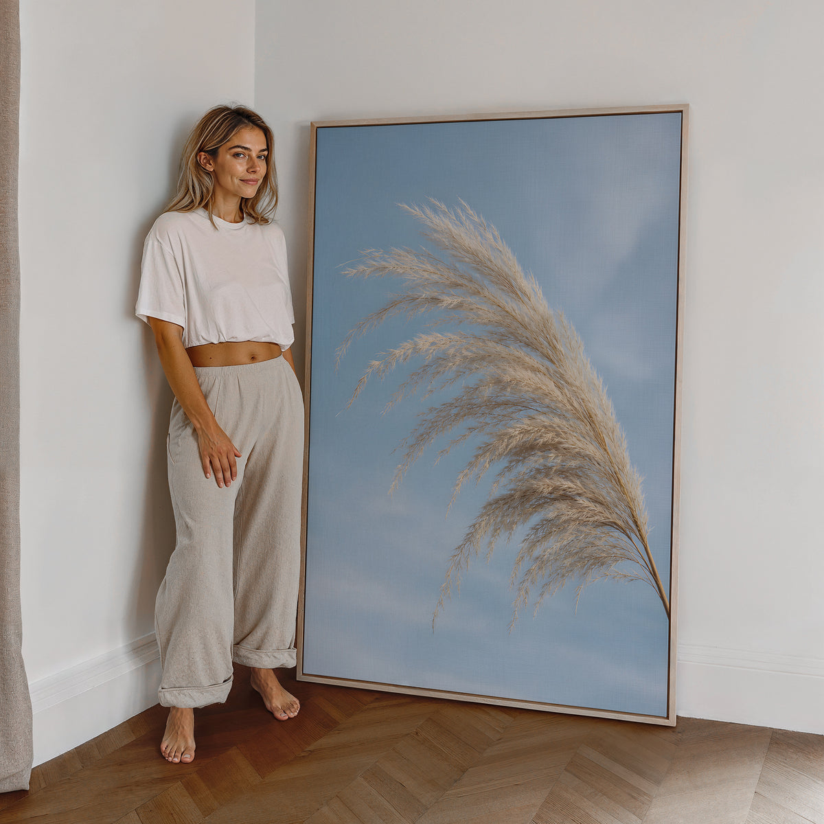 Motiv mit einem gebogenen hellen Pampaswedel vor blauem Himmel, als großes gerahmtes Wandbild neben einer Frau.