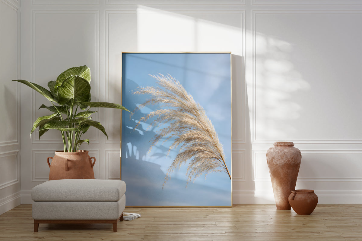 Motiv mit einem gebogenen hellen Pampaswedel vor blauem Himmel, als gerahmtes Poster neben Sessel und Zimmerpflanze.