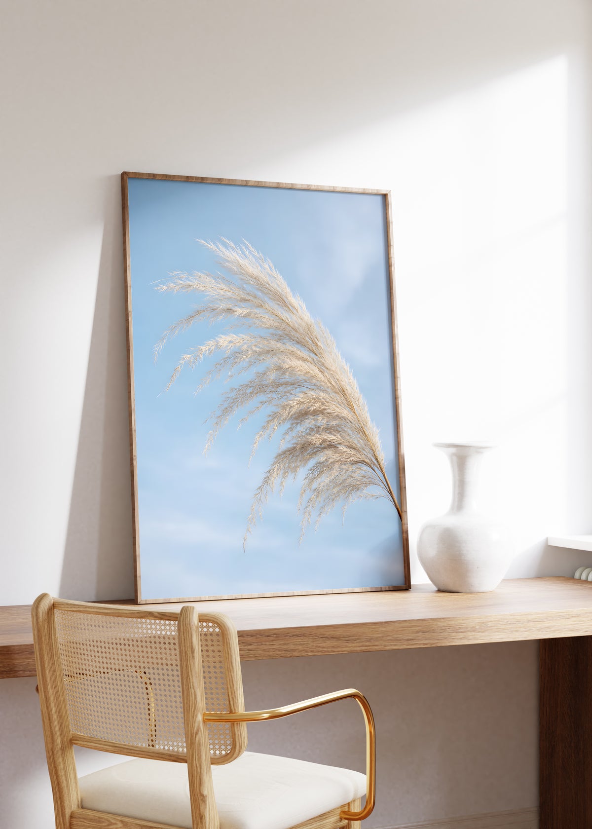 Gerahmtes Poster mit einem gebogenen hellen Pampaswedel vor blauem Himmel, auf einem Holztisch neben einer weißen Vase.