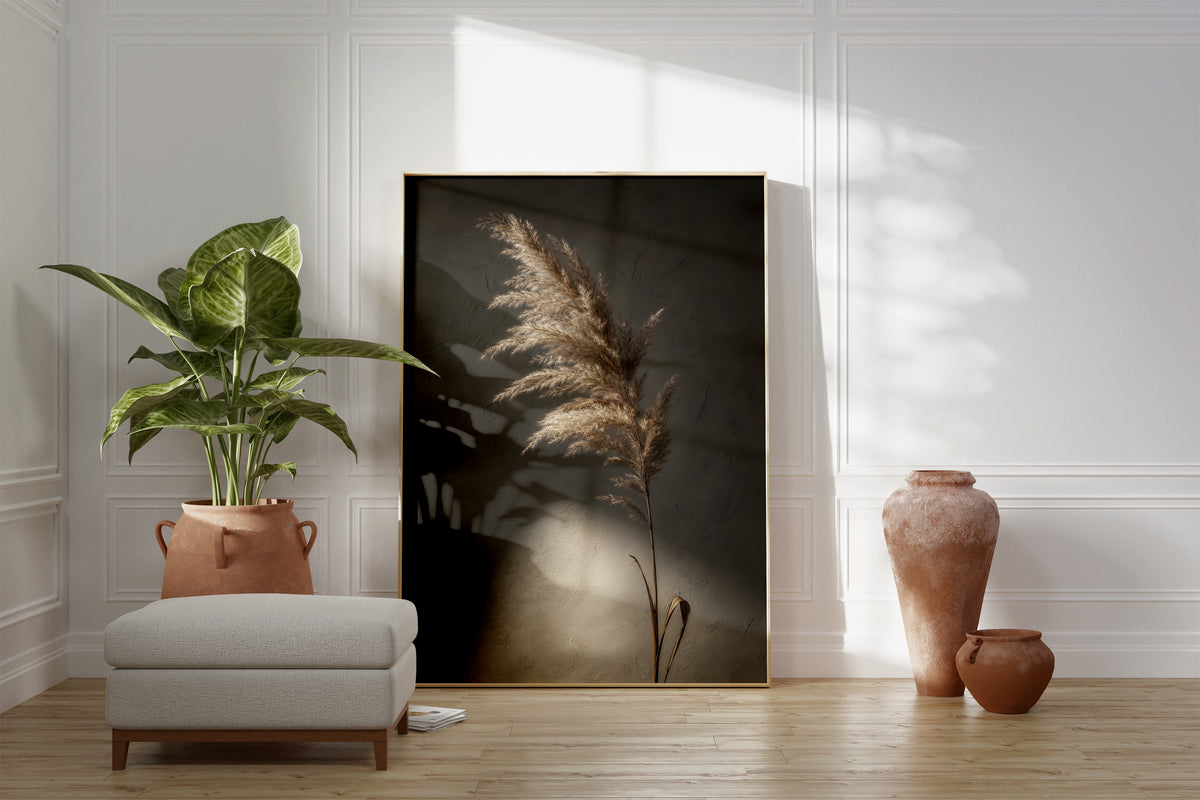 Motiv mit einem braunbeigen Pampaswedel vor dunklem Hintergrund mit einem hellen Lichtfleck, als gerahmtes Poster neben Sessel und Zimmerpflanze.