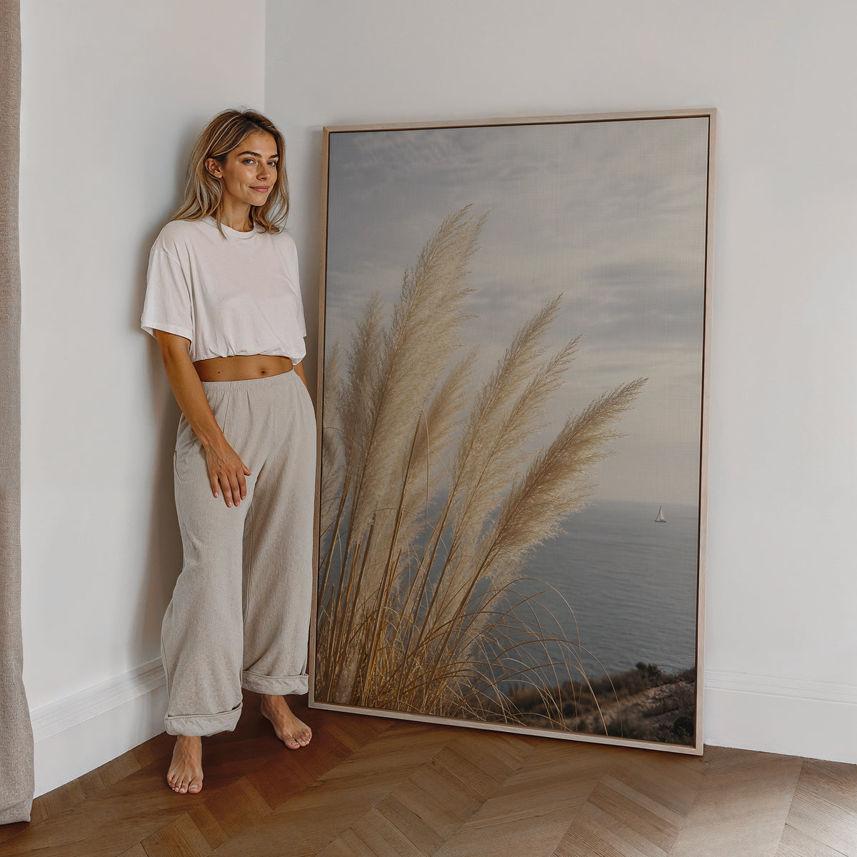 Motiv mit hohen hellen Pampaswedeln über einer ruhigen Meeresbucht, als großes gerahmtes Wandbild neben einer Frau.