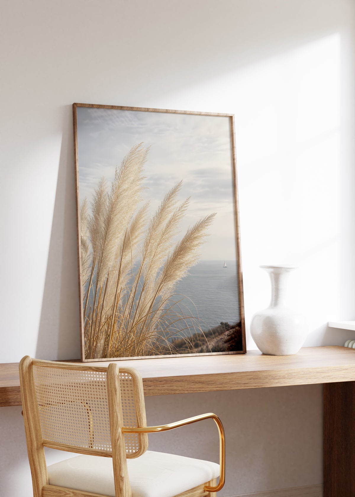 Gerahmtes Poster mit hohen hellen Pampaswedeln über einer ruhigen Meeresbucht, auf einem Holztisch neben einer weißen Vase.