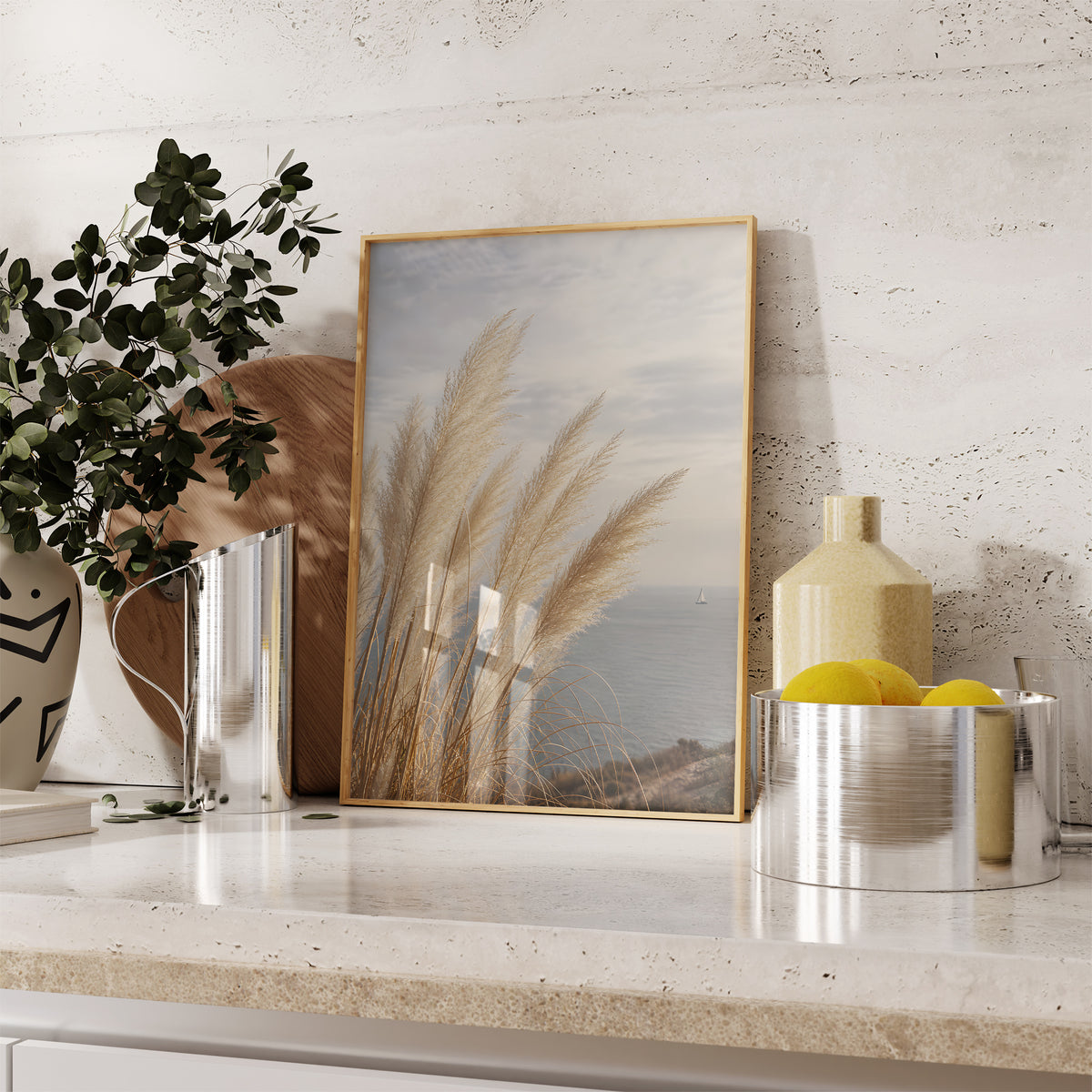 Gerahmtes Poster mit hohen hellen Pampaswedeln über einer ruhigen Meeresbucht, auf einer Küchenarbeitsplatte neben Pflanzen und Zitronen.