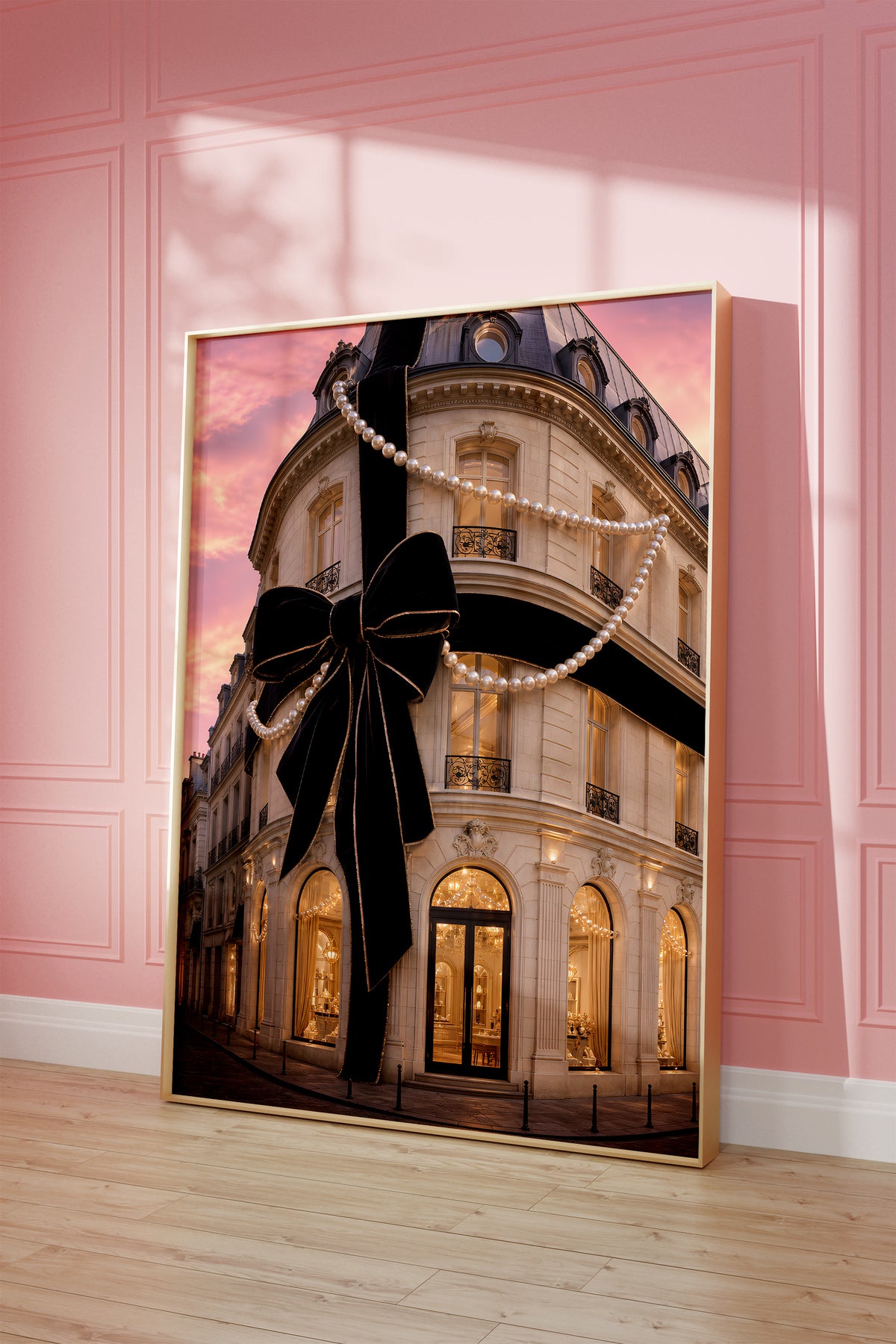 Motiv mit Pariser Hausfassade mit großer schwarzer Schleife und Perlenketten unter rosafarbenem Himmel, als gerahmtes Poster vor einer rosafarbenen Wand.