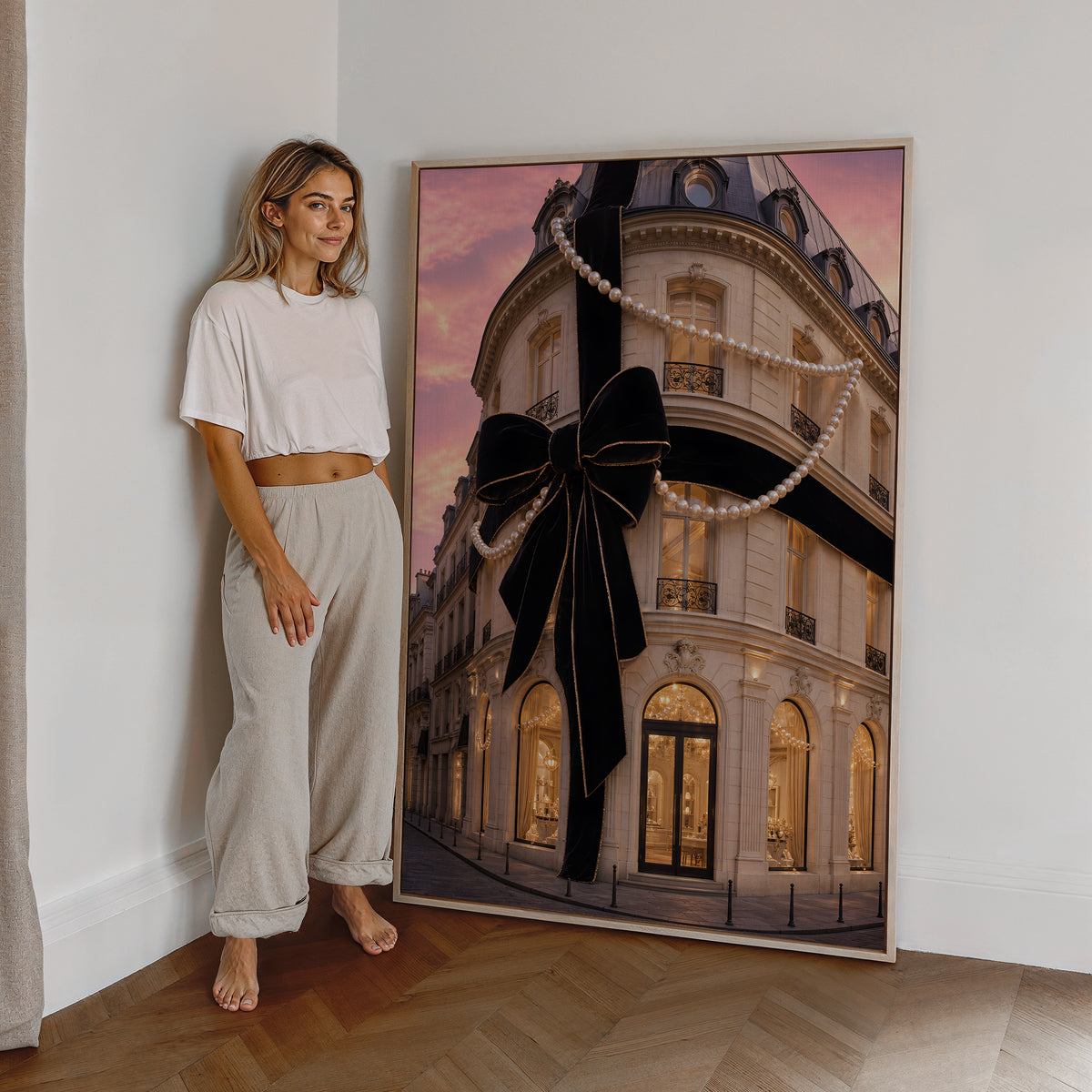 Motiv mit Pariser Hausfassade mit großer schwarzer Schleife und Perlenketten unter rosafarbenem Himmel, als großes gerahmtes Wandbild neben einer Frau.