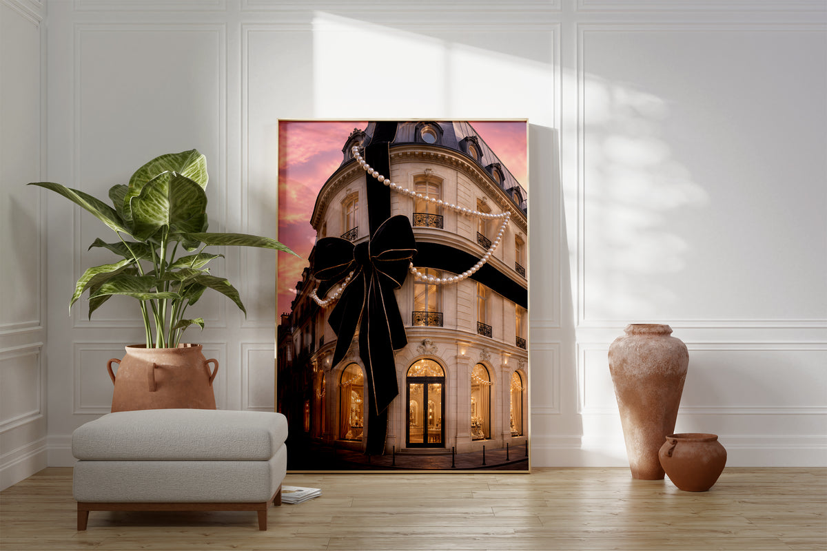 Motiv mit Pariser Hausfassade mit großer schwarzer Schleife und Perlenketten unter rosafarbenem Himmel, als gerahmtes Poster neben Sessel und Zimmerpflanze.