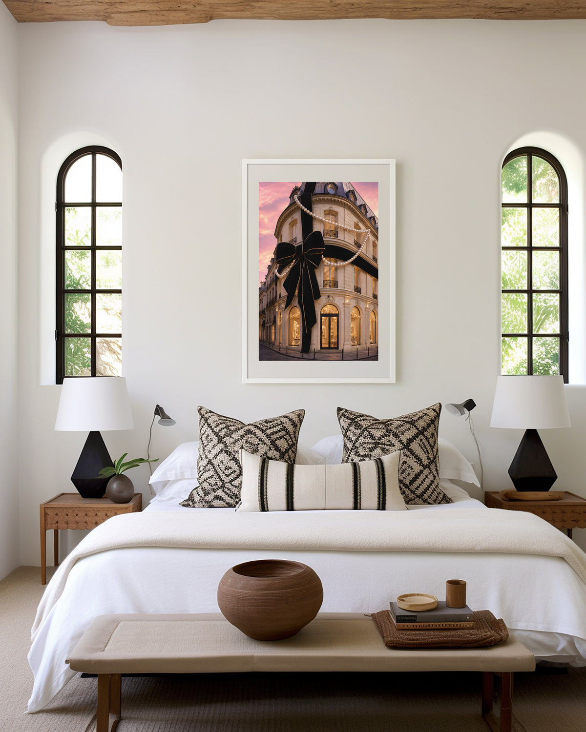 Motiv mit Pariser Hausfassade mit großer schwarzer Schleife und Perlenketten unter rosafarbenem Himmel, über einem Bett im hellen Schlafzimmer.