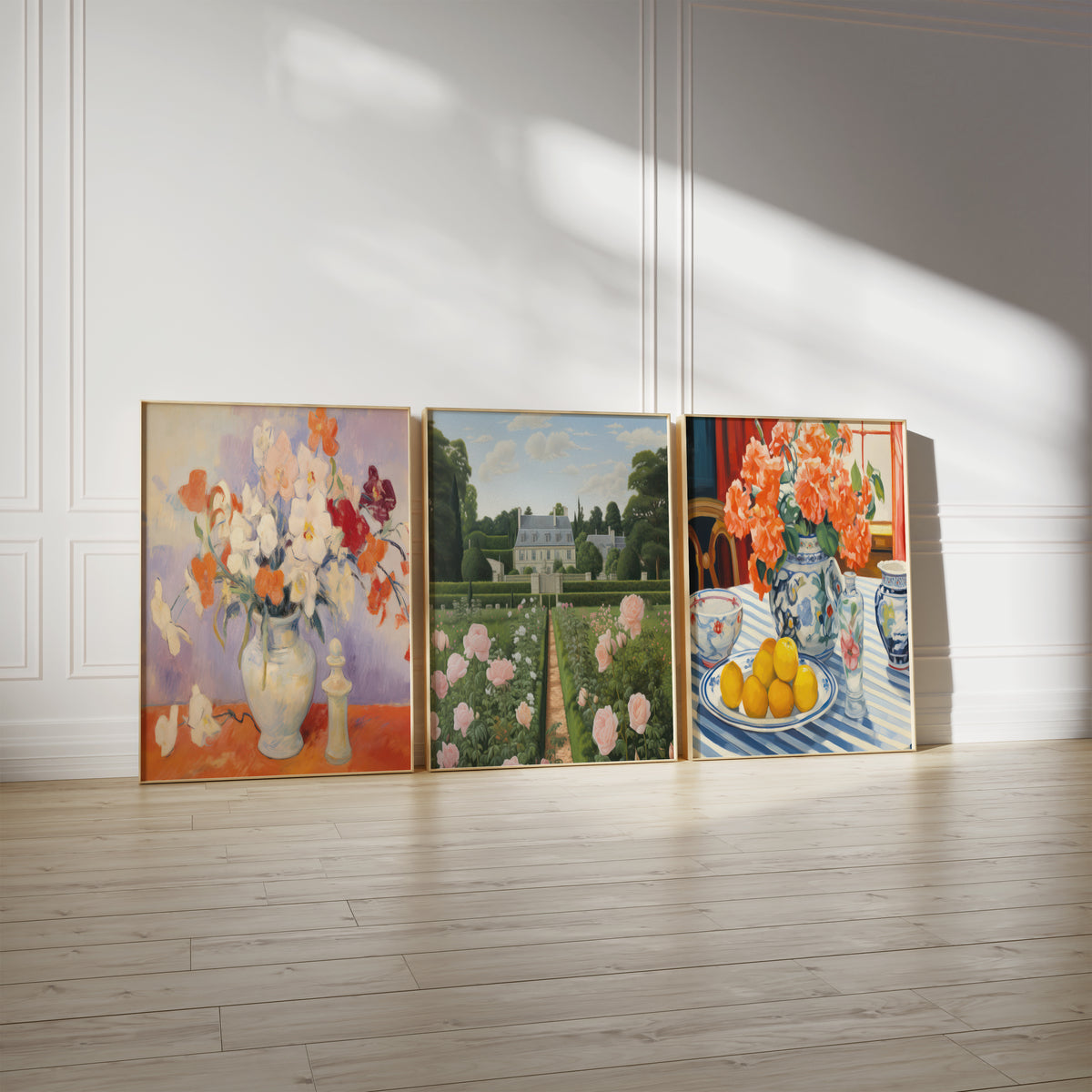 Drei gerahmte Poster an einer hellen Wand mit Zierleisten. Motive: Weiße und orange Blüten in einem hellen Krug auf einem roten Tisch; Symmetrischer Rosengarten vor einem hellen Schloss; Orangefarbene Blumen in einer blau-weißen Vase neben Zitronen.