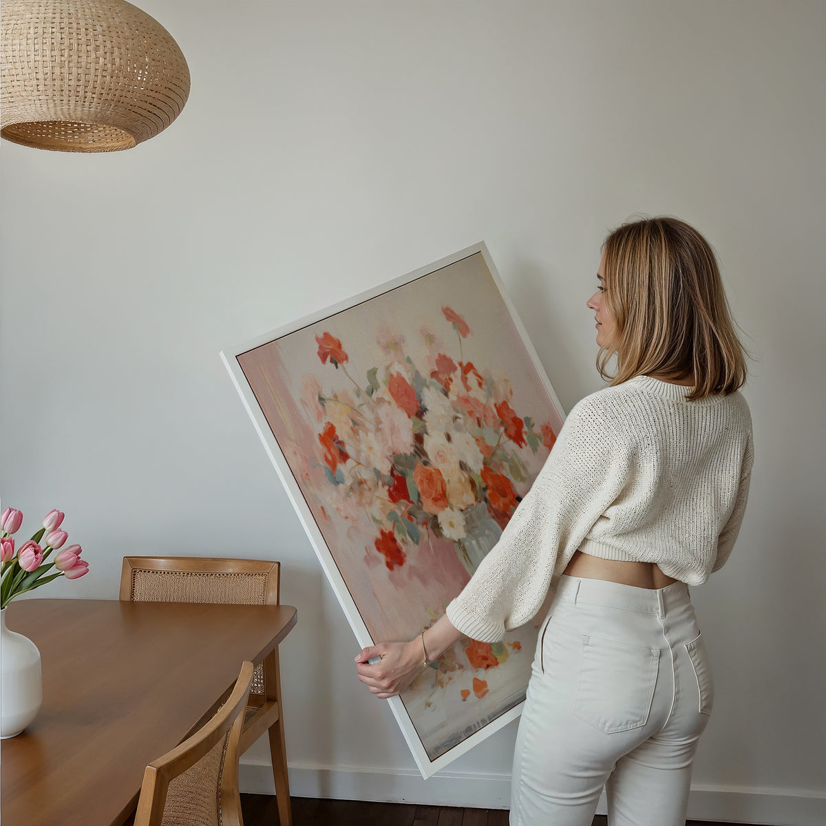 Eine Frau hält ein großes gerahmtes Poster im Essbereich mit Holzmöbeln. Motiv: Lockerer Blumenstrauß in Rosa, Rot und Weiß in einer hellen Vase.