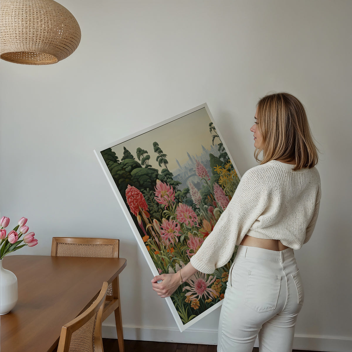 Eine Frau hält ein großes gerahmtes Poster im Essbereich mit Holzmöbeln. Motiv: Farbenfroher Blumengarten mit rosa und roten Blüten vor einer fernen Stadt.