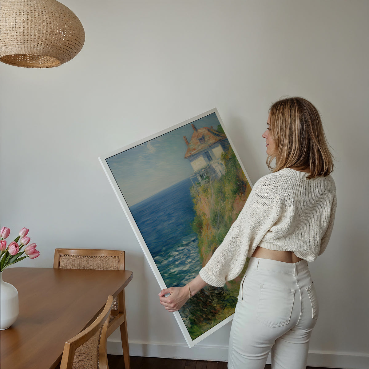 Eine Frau hält ein großes gerahmtes Poster im Essbereich mit Holzmöbeln. Motiv: Helles Haus an einer begrünten Steilküste über dem blauen Meer.
