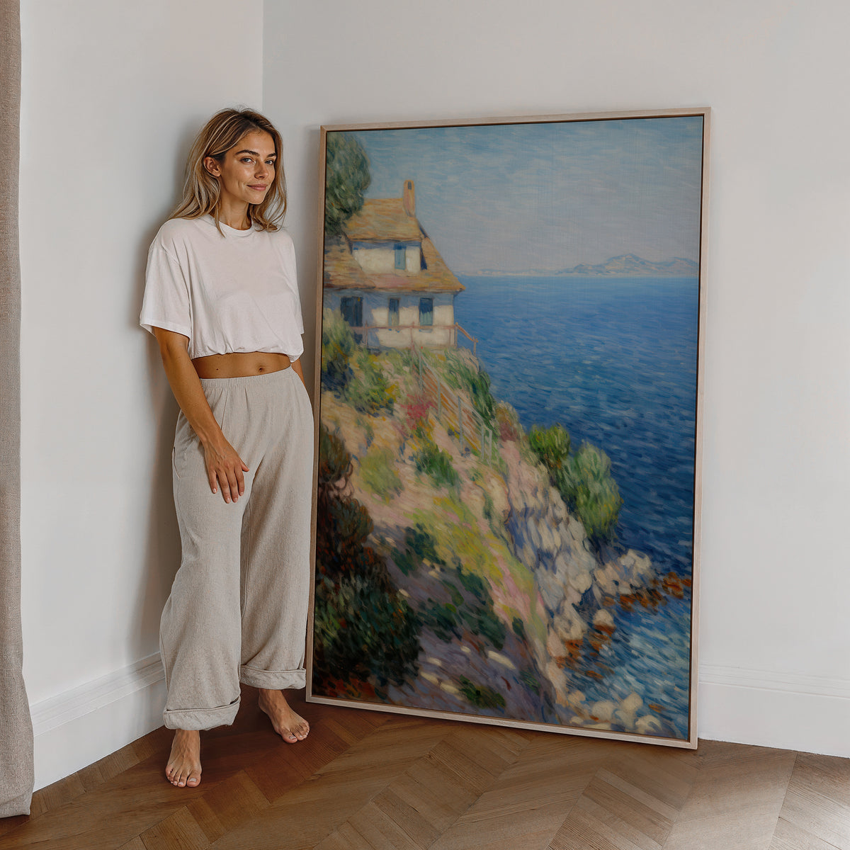 Eine Frau steht neben einem großen gerahmten Poster. Motiv: Helles Haus auf einer felsigen Küste über dem blauen Meer.
