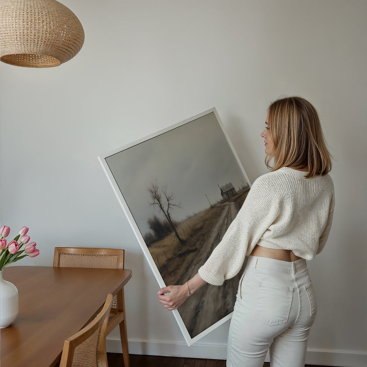 Eine Frau hält ein großes gerahmtes Poster im Essbereich mit Holzmöbeln. Motiv: Ländlicher Weg zu einem einsamen Haus unter grauem Himmel.