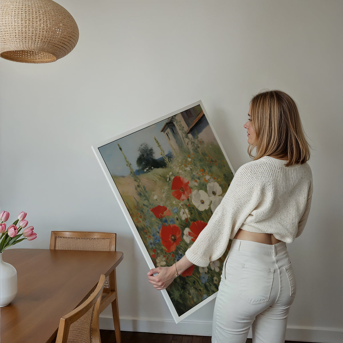 Eine Frau hält ein großes gerahmtes Poster im Essbereich mit Holzmöbeln. Motiv: Rote und weiße Mohnblumen auf einer Wiese neben einem Landhaus.