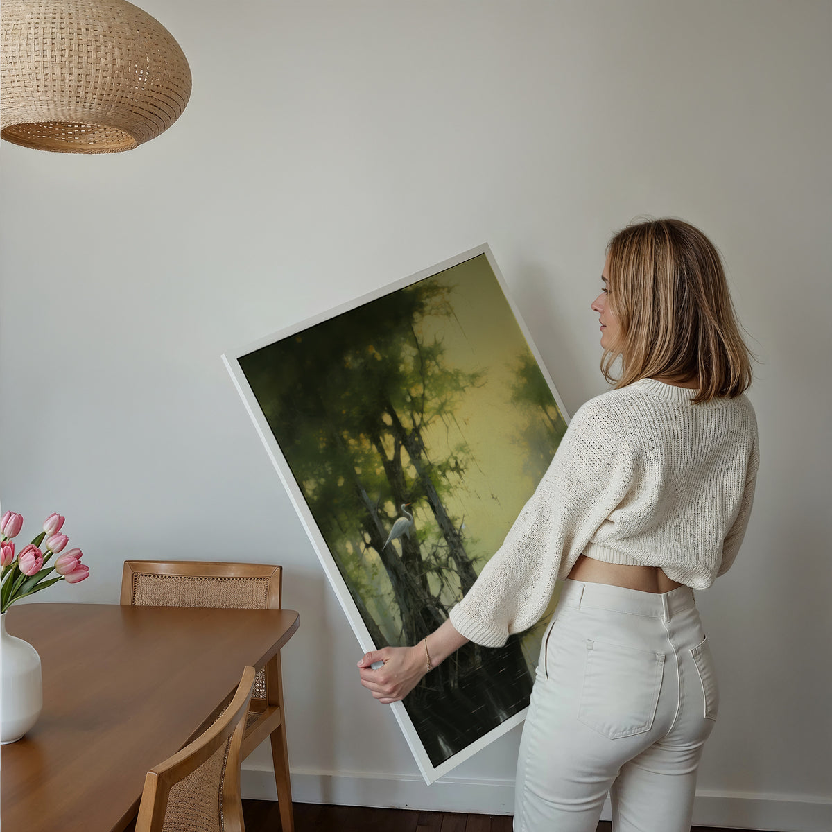 Eine Frau hält ein großes gerahmtes Poster im Essbereich mit Holzmöbeln. Motiv: Weißer Vogel zwischen Bäumen an einem nebligen Gewässer.