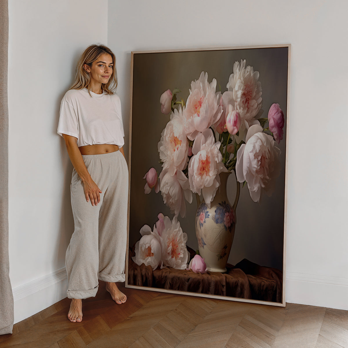 Eine Frau steht neben einem großen gerahmten Poster. Motiv: Zarte rosa Pfingstrosen in einer dekorativen Vase.
