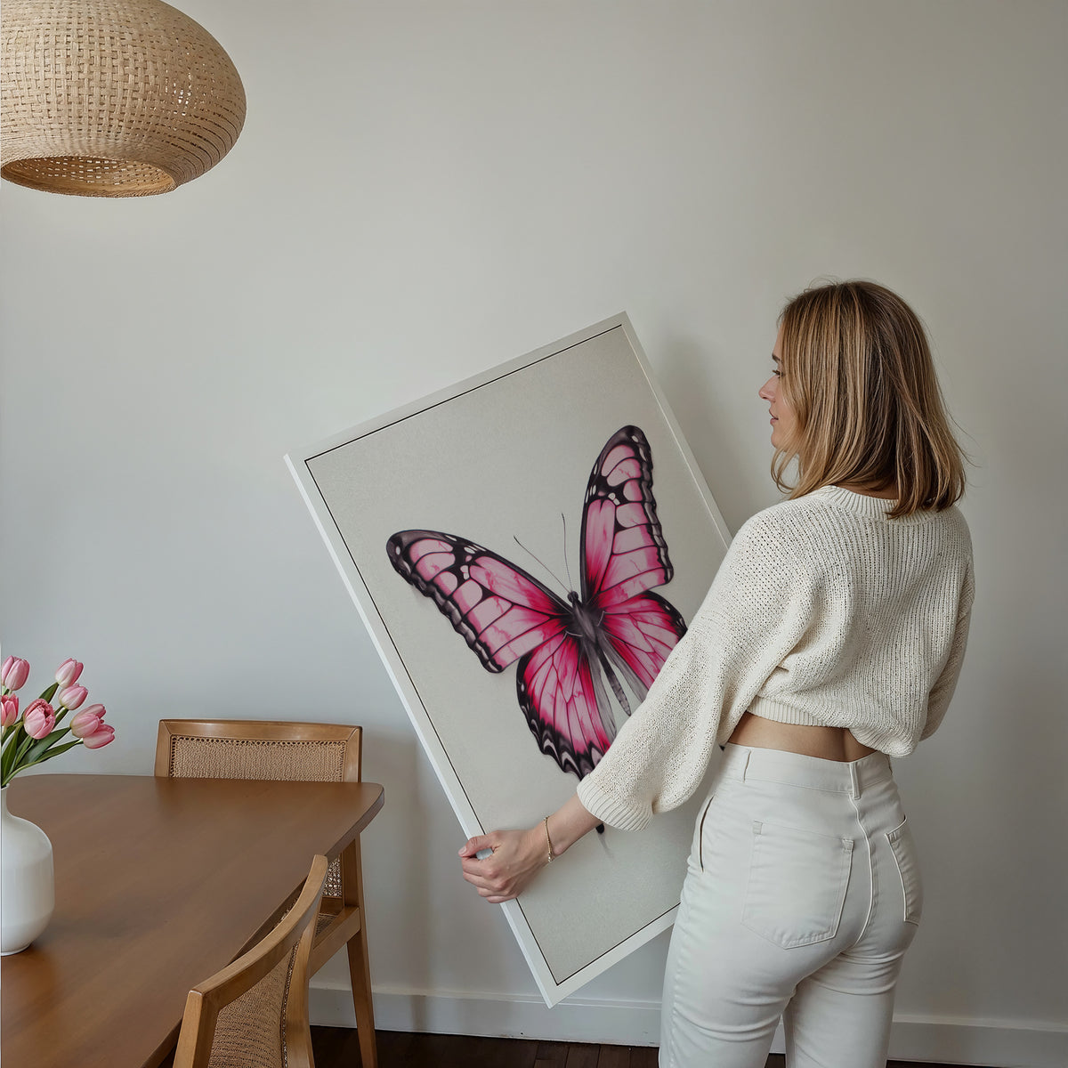 Eine Frau hält ein großes gerahmtes Poster im Essbereich mit Holzmöbeln. Motiv: Rosa Schmetterling mit schwarz umrandeten Flügeln auf hellem Hintergrund.