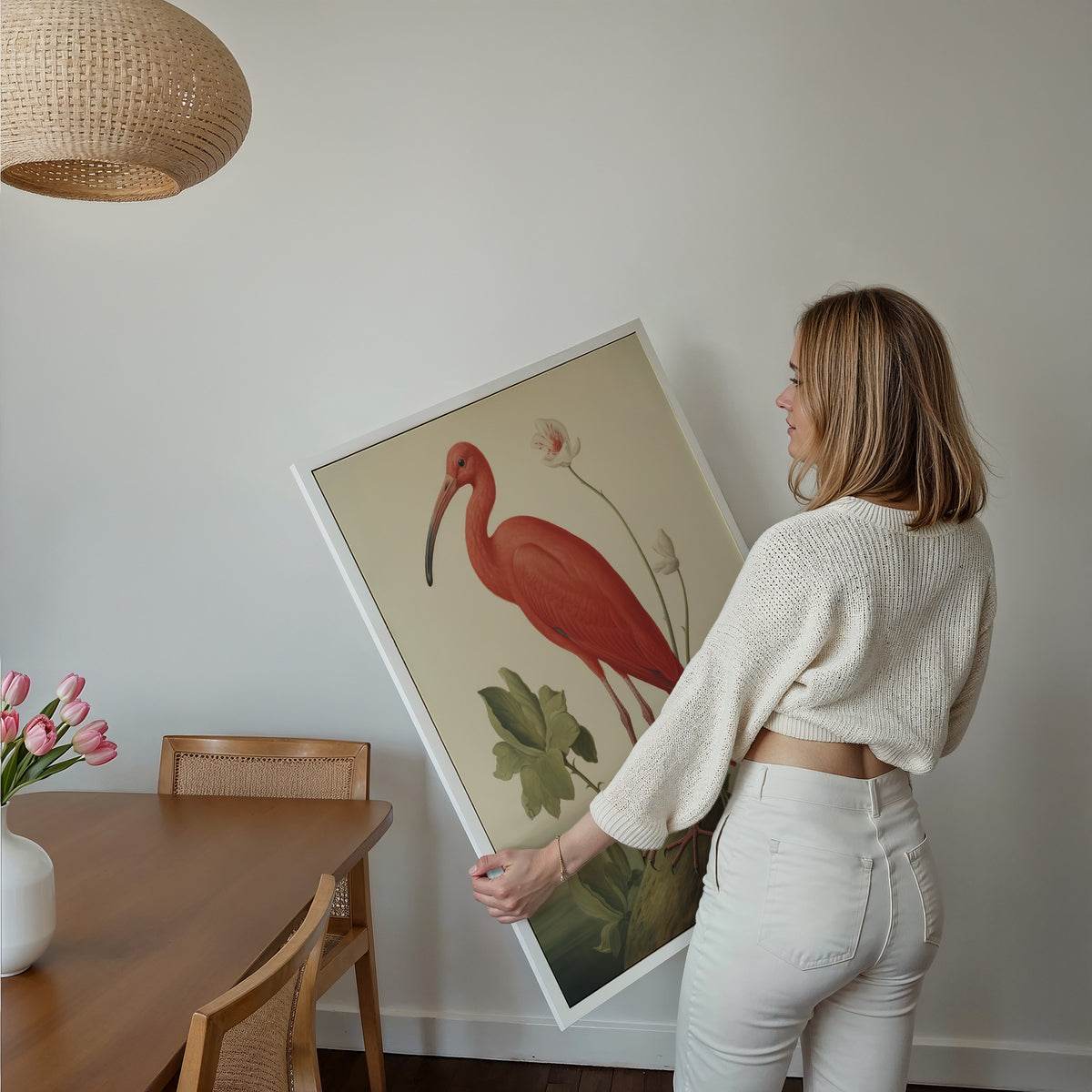 Eine Frau hält ein großes gerahmtes Poster mit einem roten Ibis im Essbereich mit Holzmöbeln und geflochtener Pendelleuchte.
