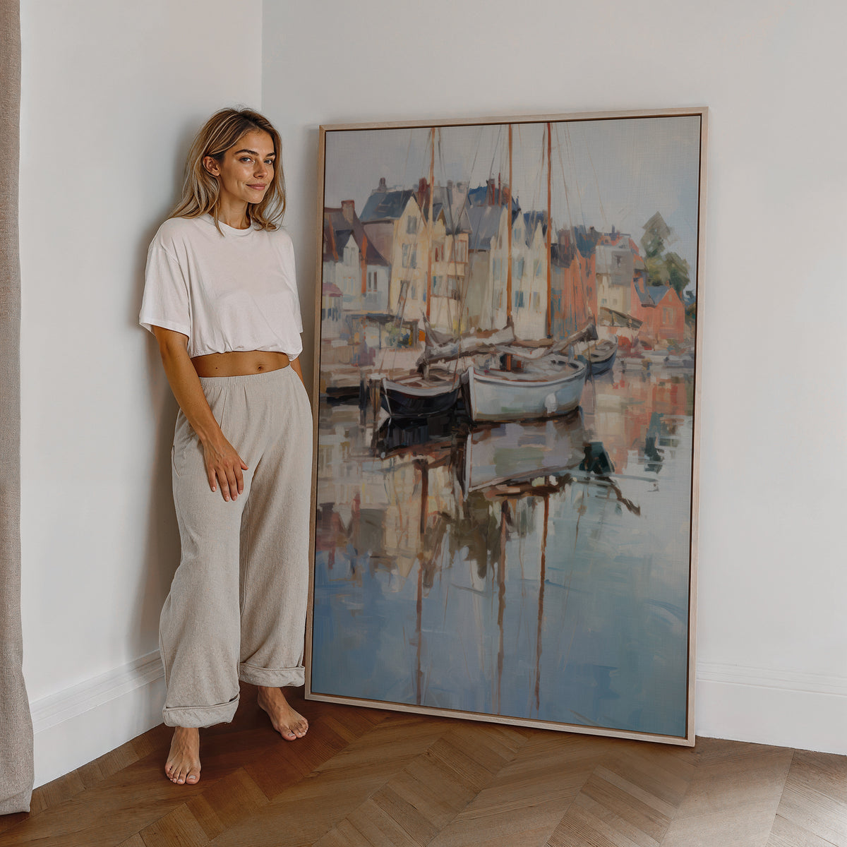 Eine Frau steht neben einem großen gerahmten Poster. Motiv: Segelboote vor pastellfarbenen Hafenhäusern mit Spiegelungen im Wasser.