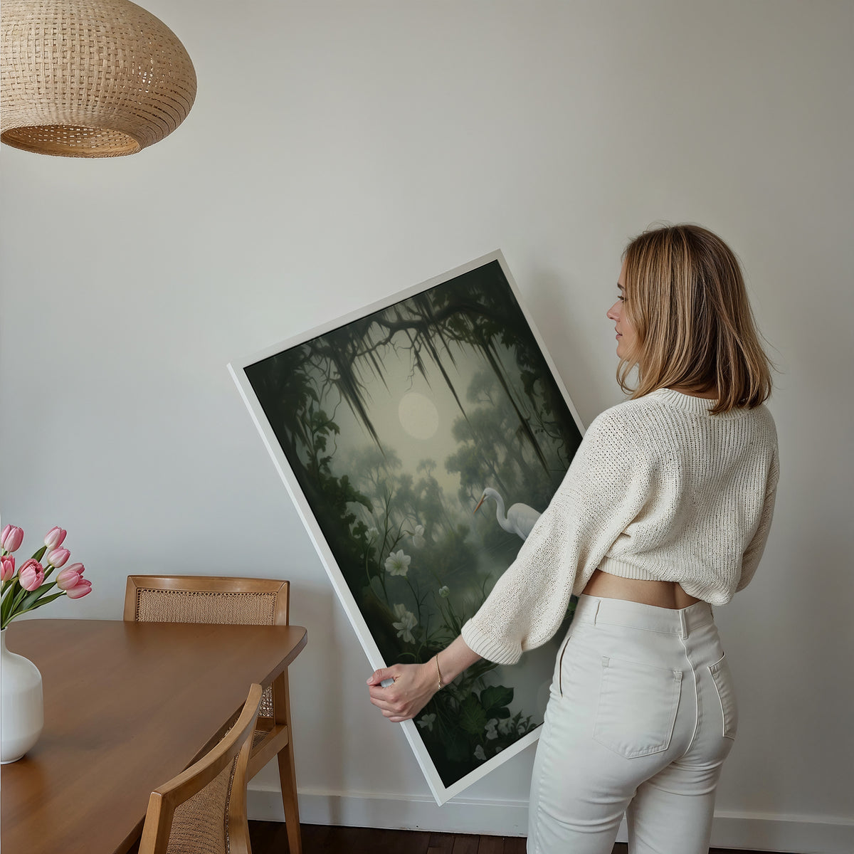 Eine Frau hält ein großes gerahmtes Poster im Essbereich mit Holzmöbeln. Motiv: Weißer Reiher an einem stillen Teich unter einem hellen Mond und dunklen Baumzweigen.