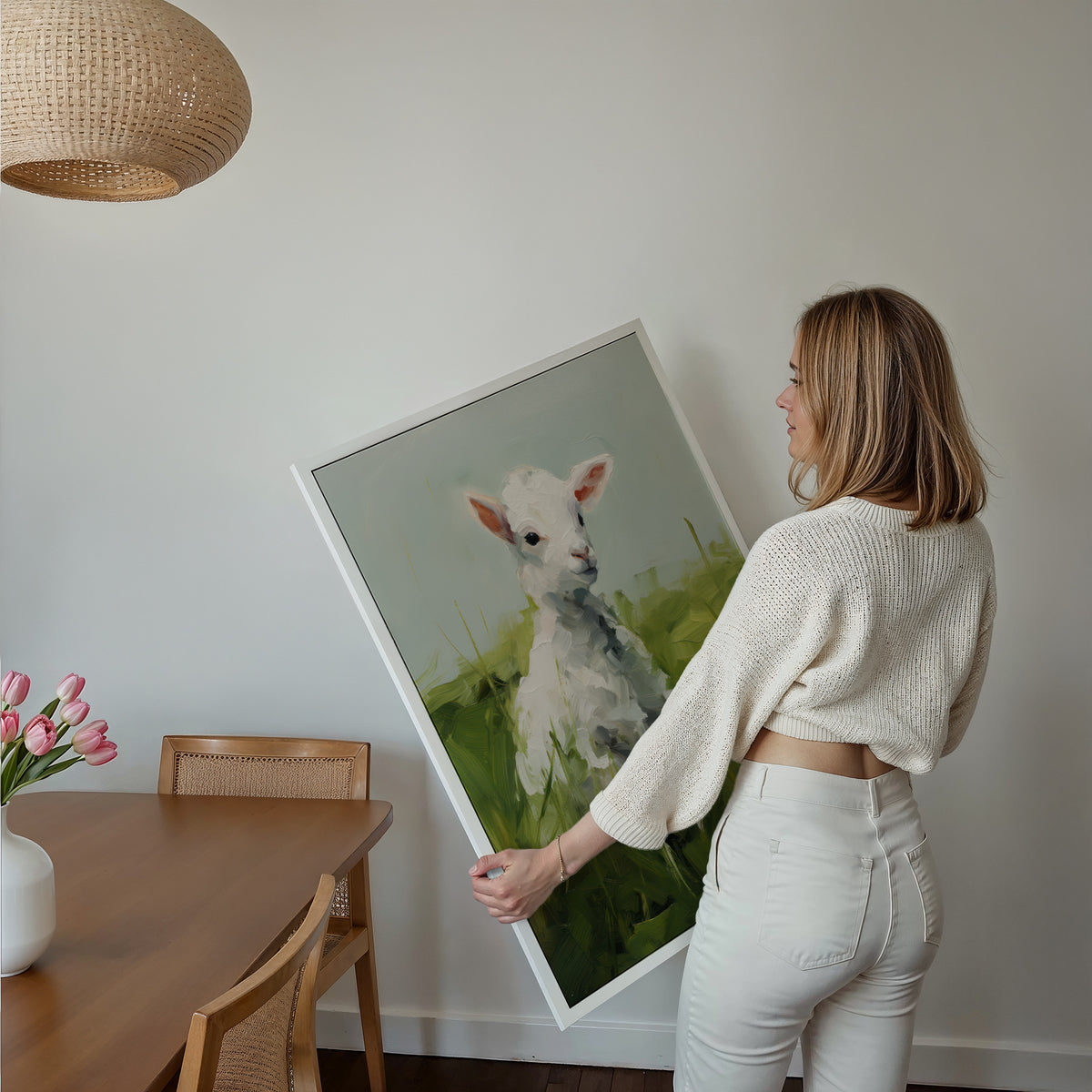 Eine Frau hält ein großes gerahmtes Poster im Essbereich mit Holzmöbeln. Motiv: Weißes Lamm auf einer grünen Wiese.