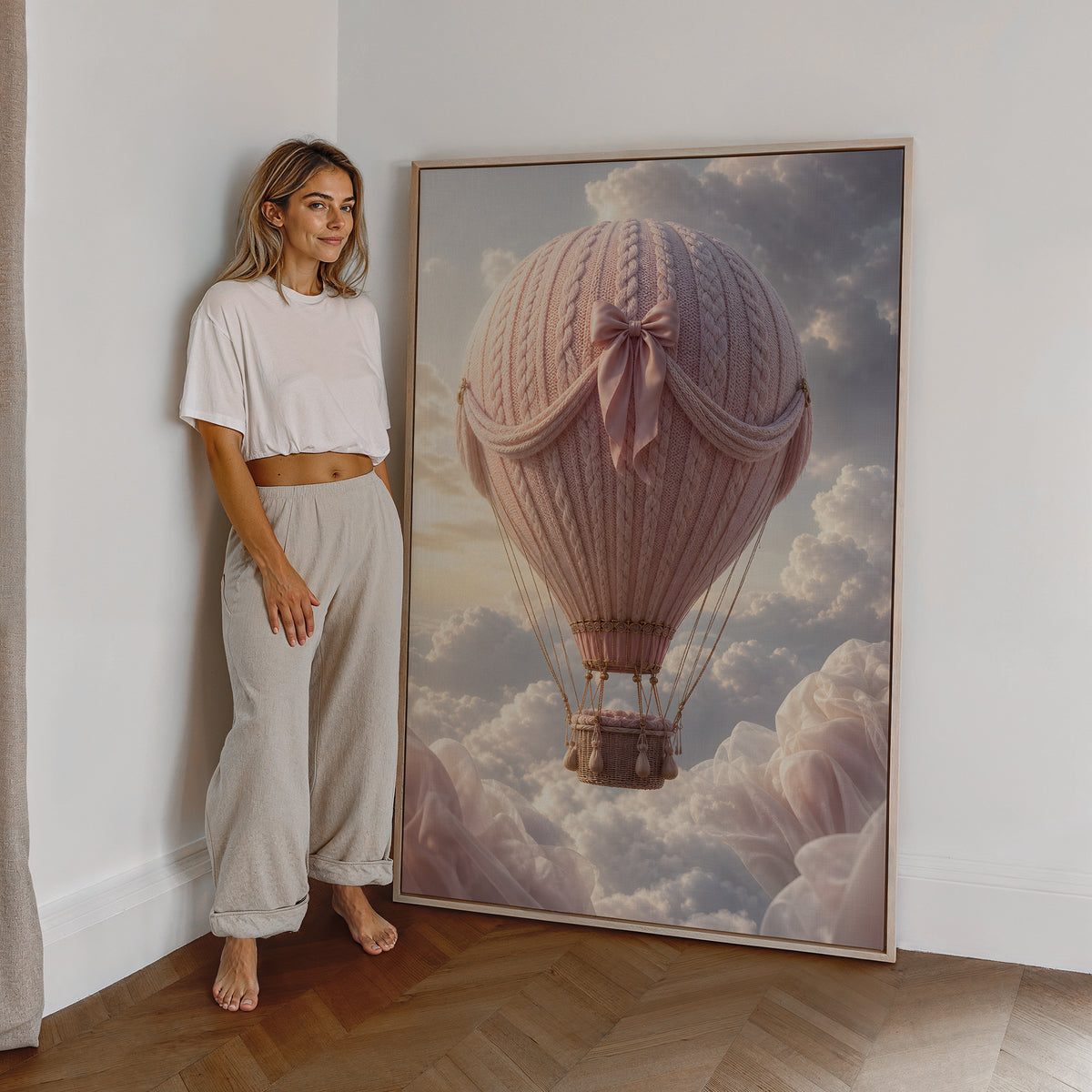 Motiv mit einem rosafarbenen Heißluftballon in Strickoptik mit Satinschleife über Wolken, als großes gerahmtes Wandbild neben einer Frau.