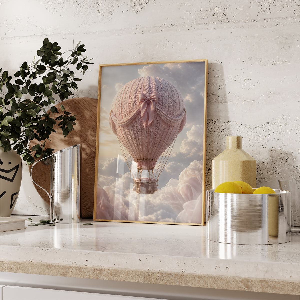 Gerahmtes Poster mit einem rosafarbenen Heißluftballon in Strickoptik mit Satinschleife über Wolken, auf einer Küchenarbeitsplatte neben Pflanzen und Zitronen.
