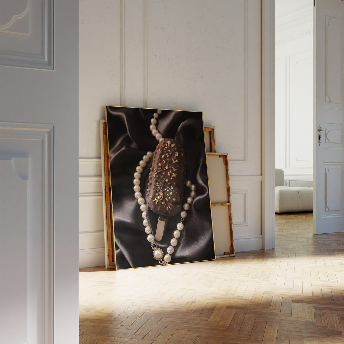 Motiv mit einem Schokoladeneis am Stiel mit Blattgold auf schwarzem Satin neben einer Perlenkette, als an die Wand gelehntes Poster neben leeren Rahmen.