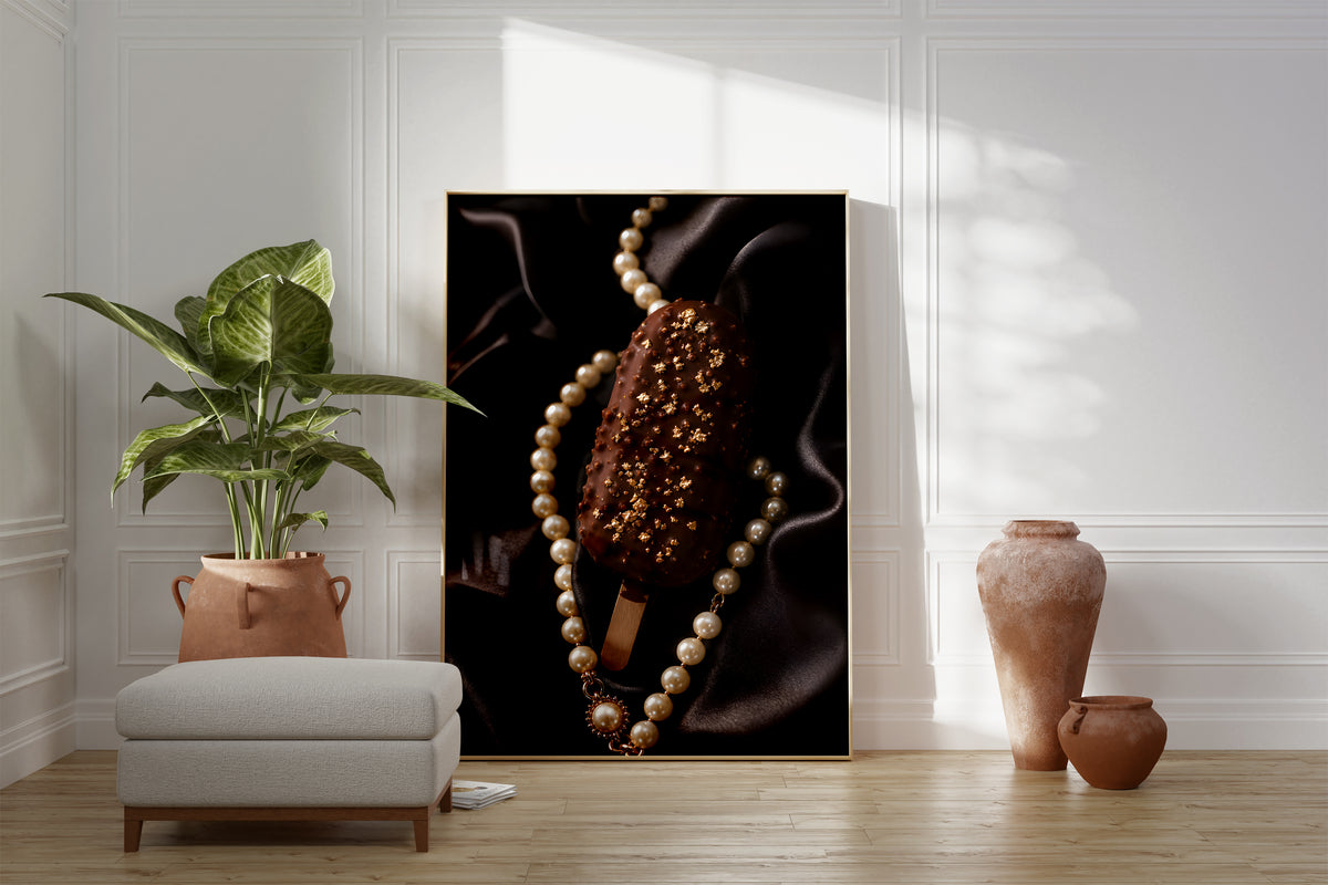 Motiv mit einem Schokoladeneis am Stiel mit Blattgold auf schwarzem Satin neben einer Perlenkette, als gerahmtes Poster neben Sessel und Zimmerpflanze.