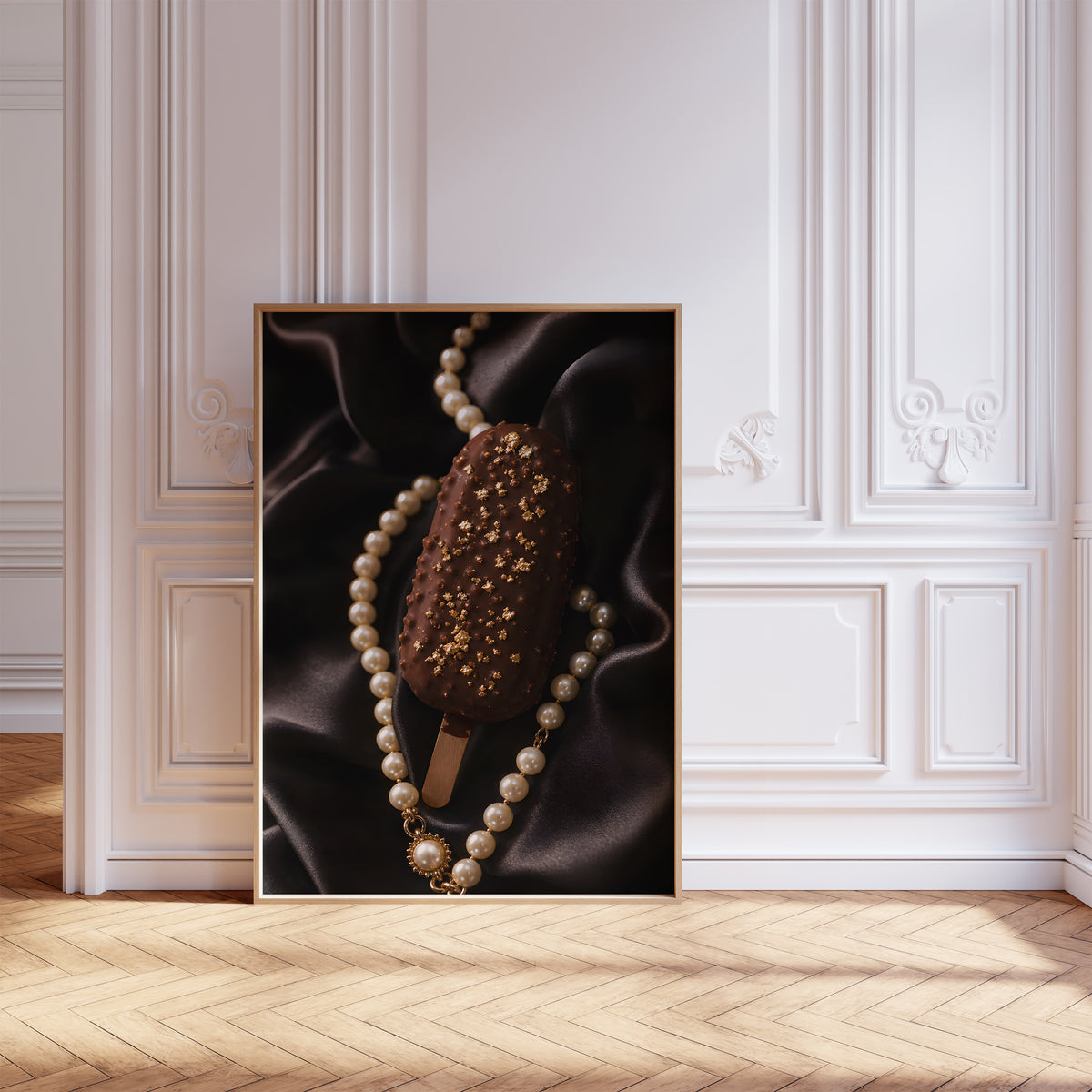 Gerahmtes Poster mit einem Schokoladeneis am Stiel mit Blattgold auf schwarzem Satin neben einer Perlenkette, vor einer weißen Wand mit Zierleisten.