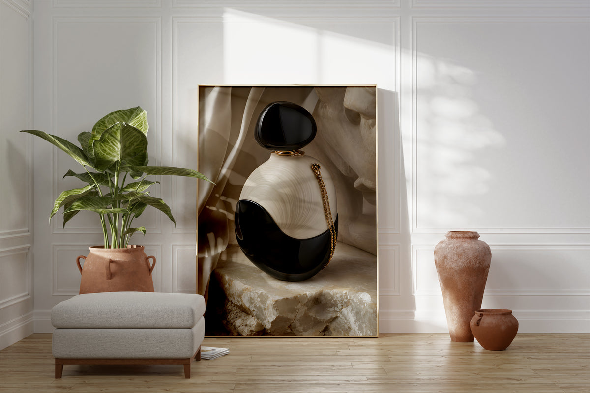 Motiv mit einem schwarz-weißen Parfumflakon mit einer goldenen Kette auf hellem Stein, als gerahmtes Poster neben Sessel und Zimmerpflanze.