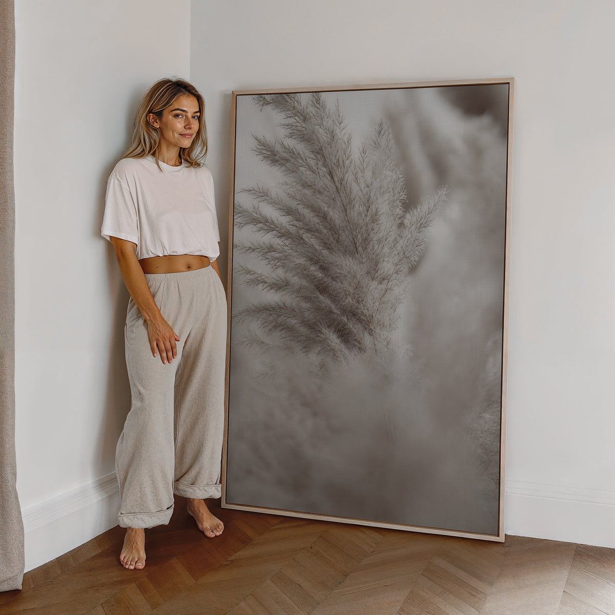 Motiv mit einem silbergrauen Pampaswedel vor weich verschwommenem Hintergrund, als großes gerahmtes Wandbild neben einer Frau.