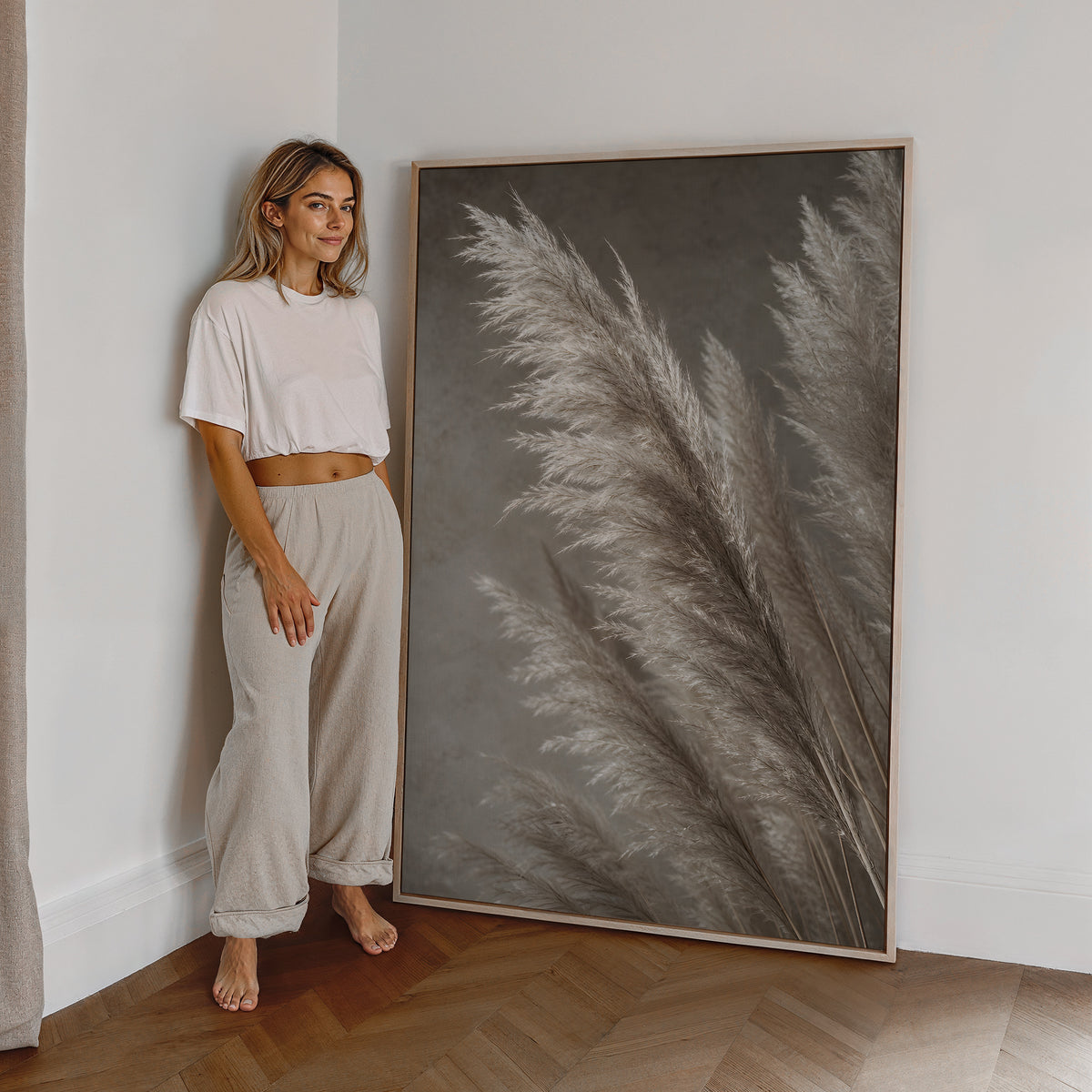 Motiv mit silbrig hellen Pampaswedeln vor grauem Hintergrund, als großes gerahmtes Wandbild neben einer Frau.