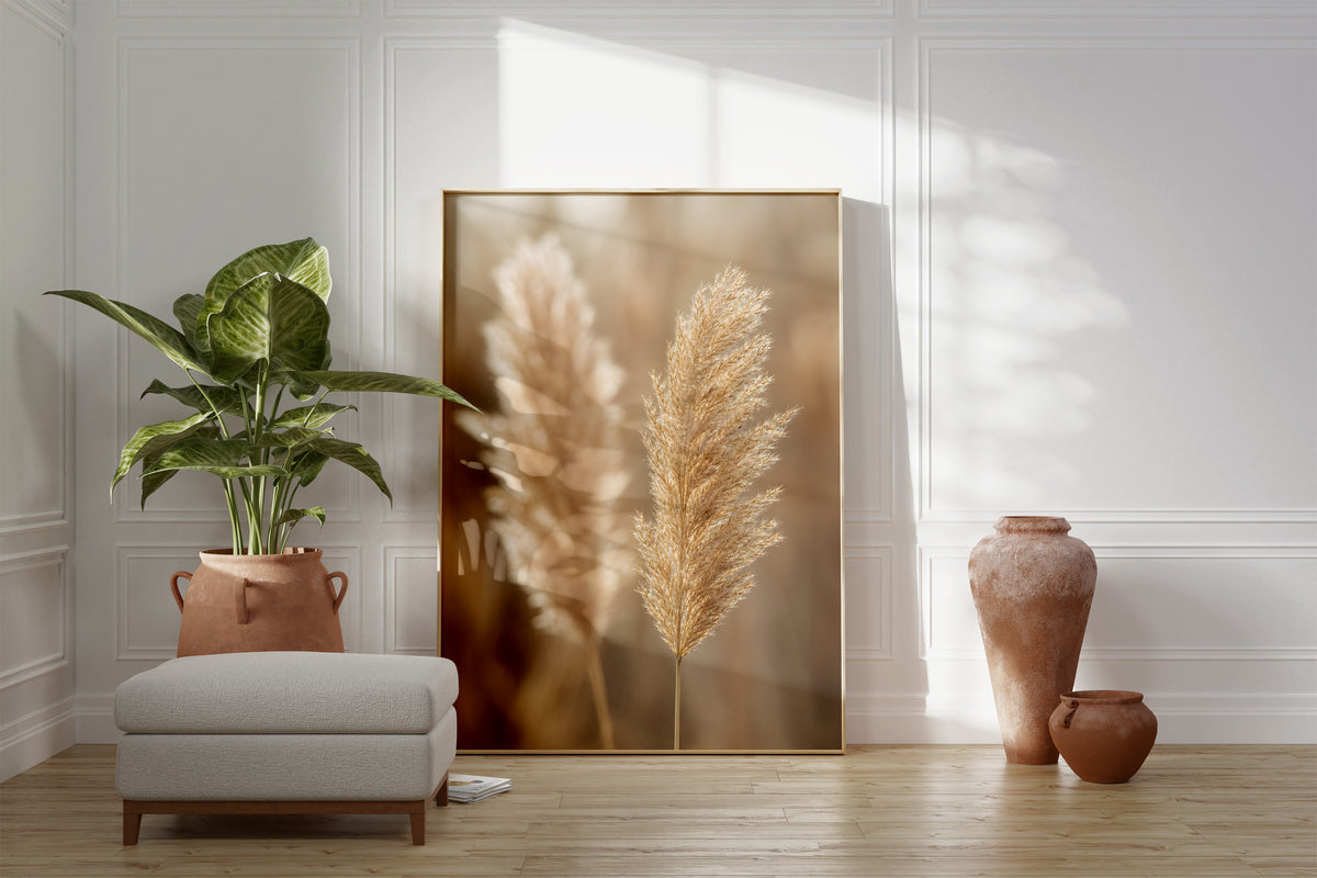 Motiv mit zwei hellbraunen Pampaswedeln vor weich verschwommenem warmbeigem Hintergrund, als gerahmtes Poster neben Sessel und Zimmerpflanze.