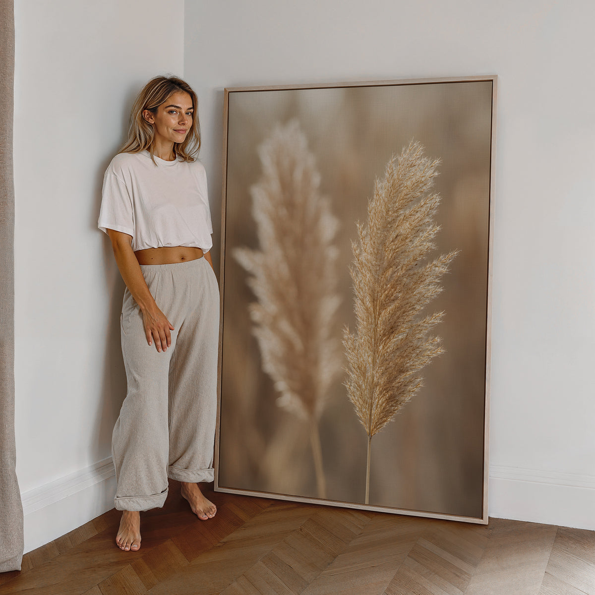 Motiv mit zwei hellbraunen Pampaswedeln vor weich verschwommenem warmbeigem Hintergrund, als großes gerahmtes Wandbild neben einer Frau.