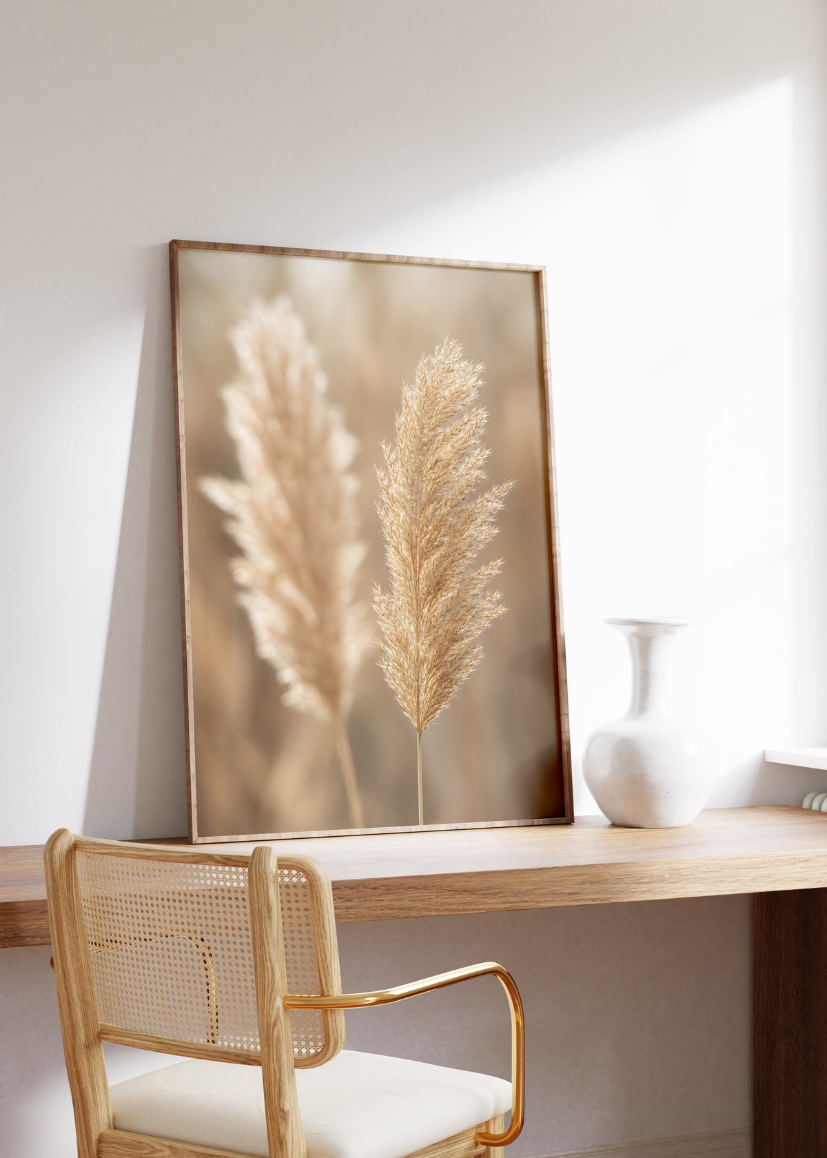 Gerahmtes Poster mit zwei hellbraunen Pampaswedeln vor weich verschwommenem warmbeigem Hintergrund, auf einem Holztisch neben einer weißen Vase.
