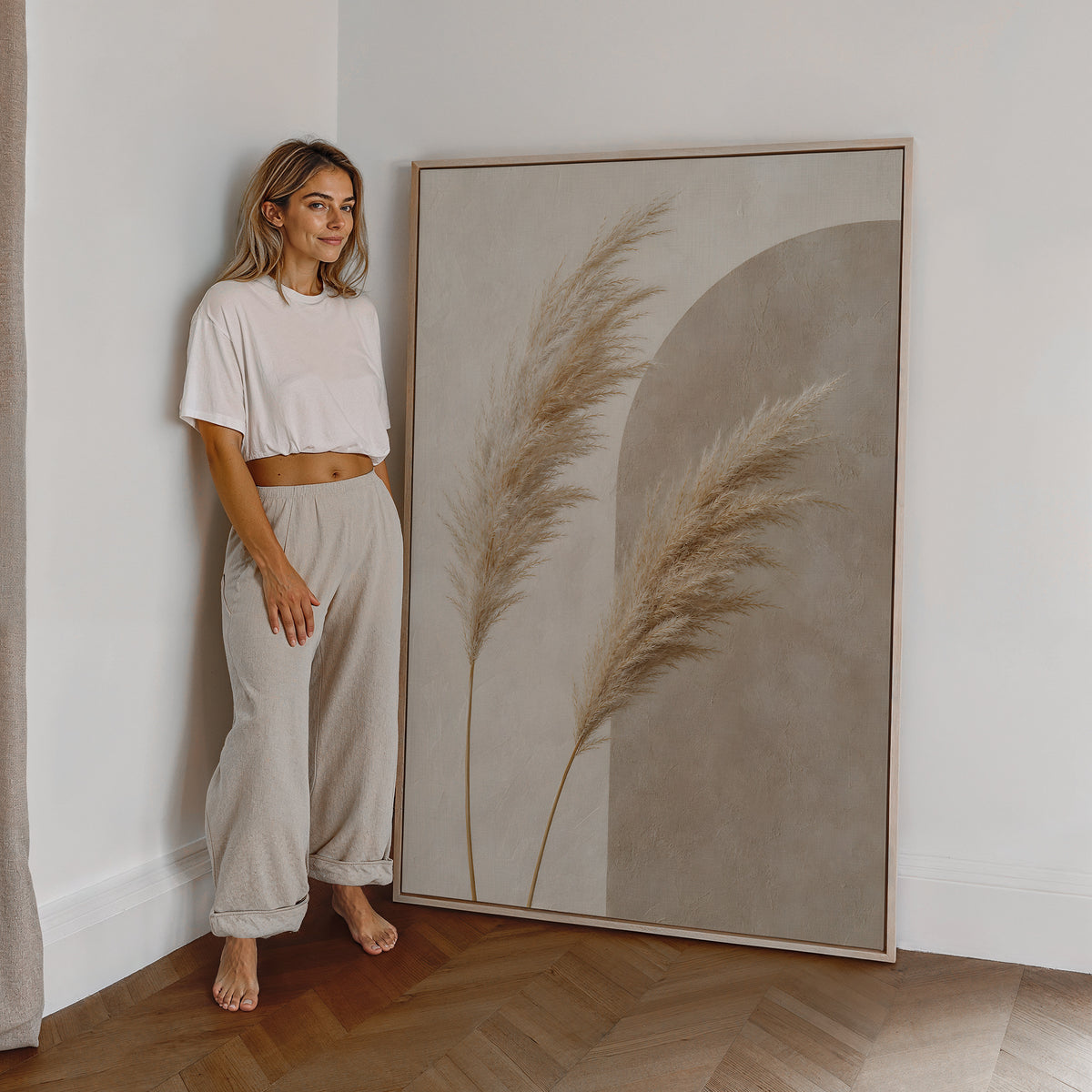 Motiv mit zwei Pampaswedeln vor einer graubeigen Bogenform, als großes gerahmtes Wandbild neben einer Frau.
