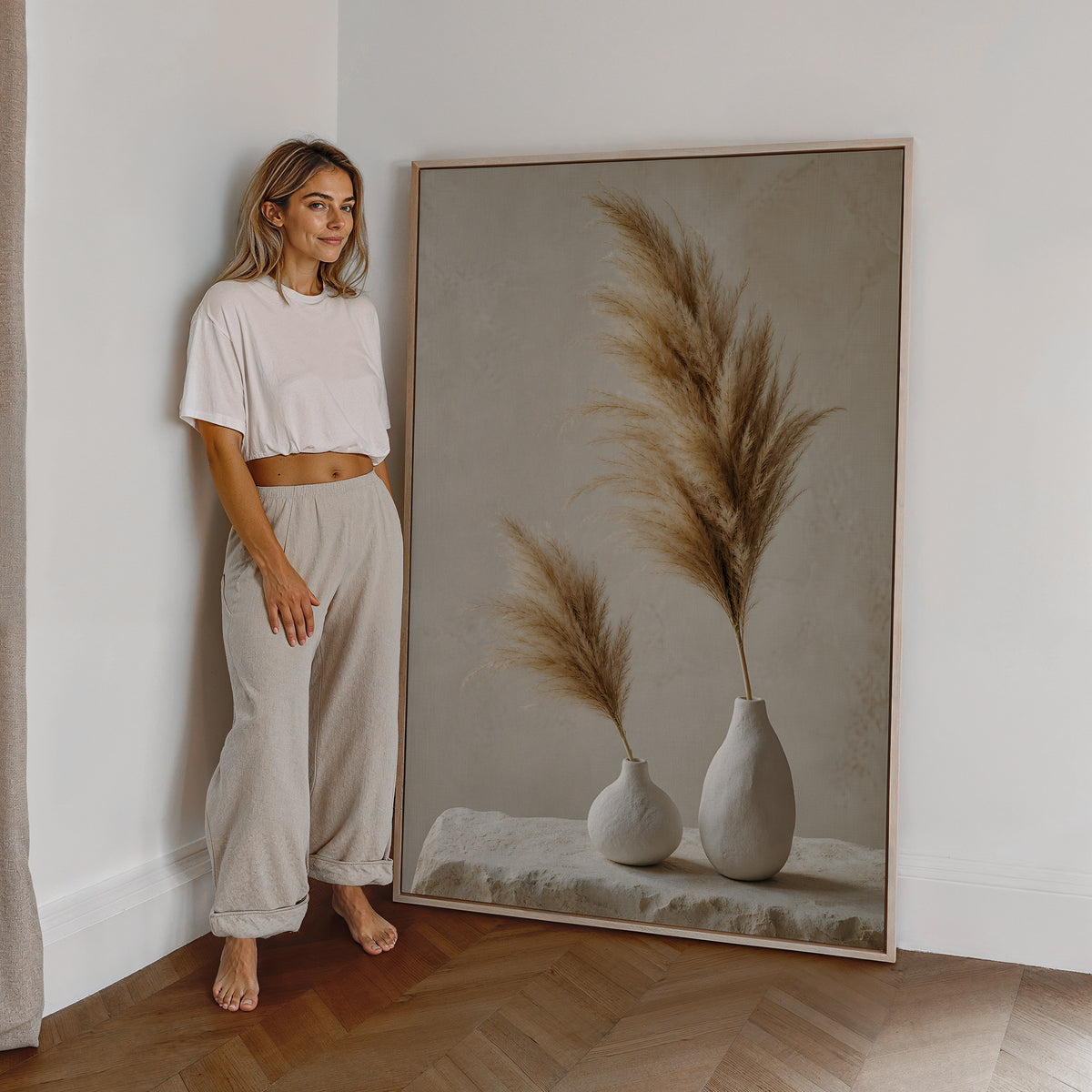 Motiv mit zwei weißen Vasen mit unterschiedlich hohen braunbeigen Pampaswedeln, als großes gerahmtes Wandbild neben einer Frau.